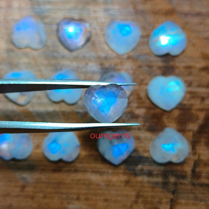May include: A collection of heart-shaped moonstone gemstones with a blue iridescent glow. One stone is held by tweezers, showcasing its facets. The word "ouregems" is visible in the image. The gemstones are arranged on a wooden surface.