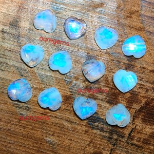 May include: A collection of heart-shaped moonstone gemstones. The stones display a range of blue and white hues, with a shimmering, iridescent quality. The word "ouregems" is printed on the surface of the image.