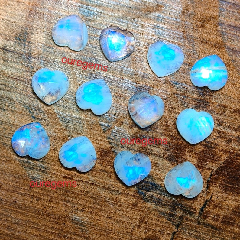 May include: A collection of heart-shaped moonstone gemstones. The stones display a range of blue and white hues, with a shimmering, iridescent quality. The word "ouregems" is printed on the surface of the image.
