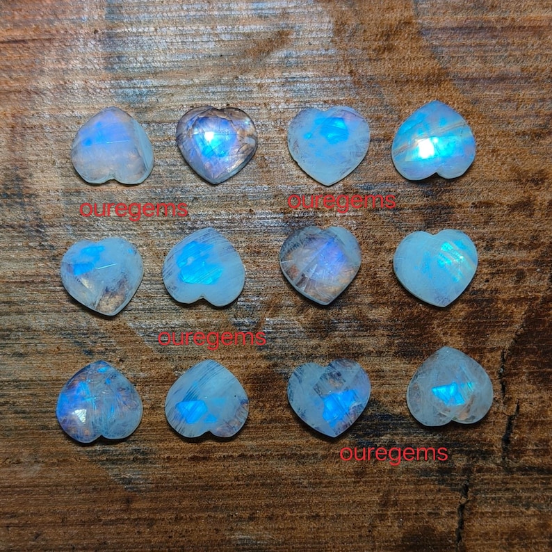 May include: A collection of heart-shaped moonstone gemstones. The stones have a translucent, milky white base with iridescent blue flashes. The gemstones are arranged on a wooden surface, with the text "puregems" visible.