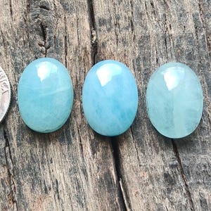 Può includere: Tre gemme ovali azzurro chiaro sono esposte su una superficie di legno stagionato accanto a una moneta. Le gemme hanno un aspetto lucido e traslucido e variano leggermente di colore. Le gemme sono probabilmente acquamarine.