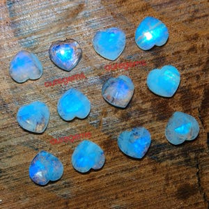 May include: A collection of heart-shaped moonstone gemstones. The stones display a range of blue and white hues, with a shimmering, iridescent quality. The gemstones are arranged on a dark wooden surface, showcasing their unique patterns and colors.