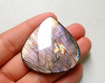 May include: A teardrop-shaped labradorite gemstone with a polished surface. The stone displays a mix of colors, including purple, gray, and hints of blue and gold. The gemstone is held in a hand.
