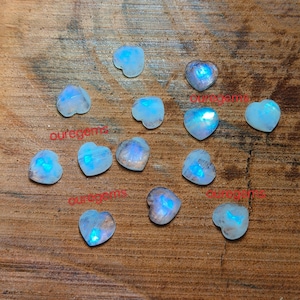 May include: A collection of heart-shaped moonstone gemstones with a blue iridescent glow. The gemstones are scattered on a wooden surface. The word "ouregems" is printed on the surface in red.