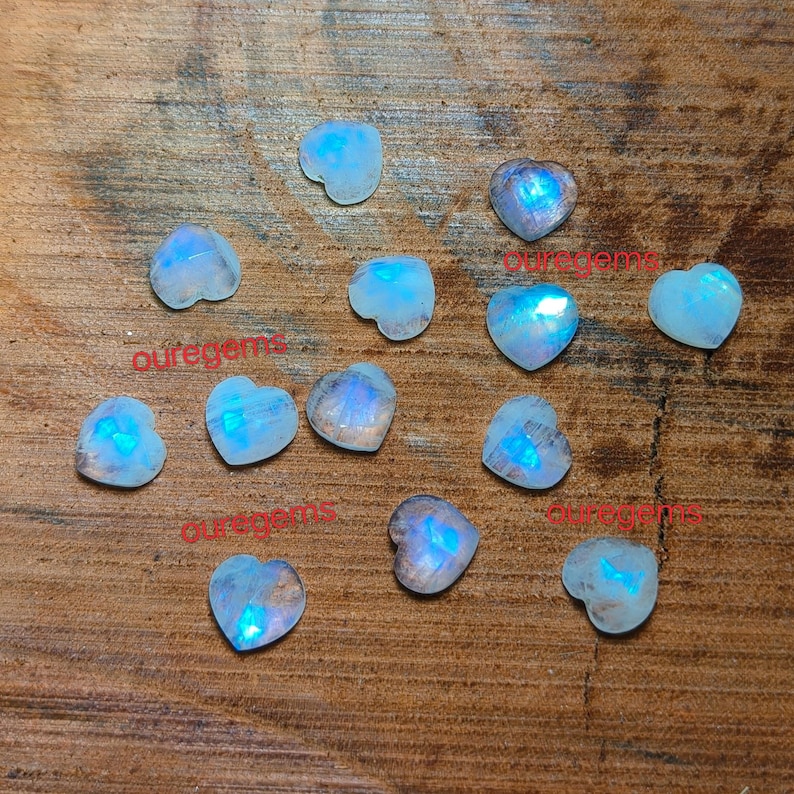 May include: A collection of heart-shaped moonstone gemstones with a blue iridescent glow. The gemstones are scattered on a wooden surface. The word "ouregems" is printed on the surface in red.