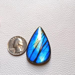 Pode incluir: Uma pedra preciosa de labradorita polida em forma de gota, com iridescência azul e verde vibrante. A pedra está ao lado de uma moeda para comparação de tamanho. A pedra tem estrias azuis escuras e pretas.