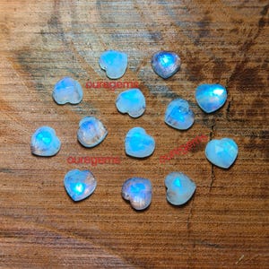 May include: A collection of heart-shaped moonstone gemstones with a blue iridescent glow. The gemstones are scattered on a wooden surface. The text "oure gems" is visible on the surface.