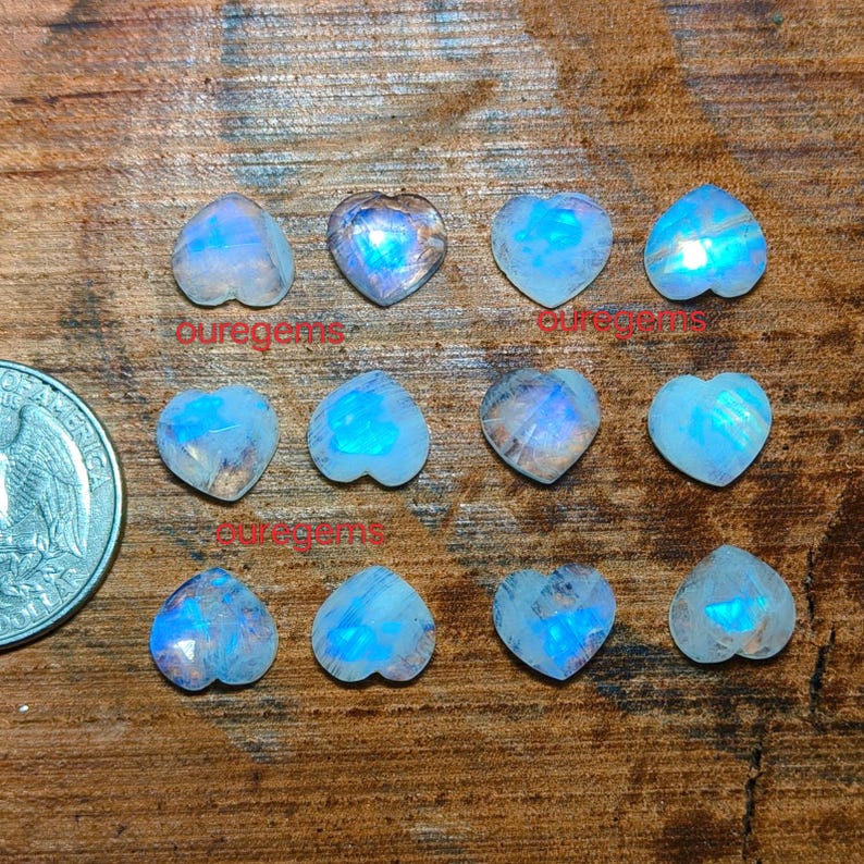 May include: A collection of heart-shaped moonstone gemstones with iridescent blue and white hues. The gemstones are arranged on a wooden surface, with a US quarter coin for size comparison. The text "puregems" is visible.