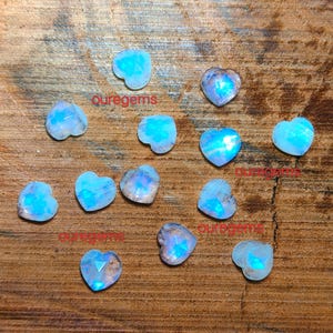 May include: A collection of heart-shaped gemstones with a blue and white iridescent glow. The gemstones are scattered on a wooden surface. The text "quregems" is visible in multiple places.