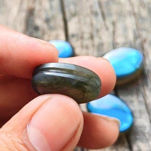 May include: A smooth, oval-shaped labradorite gemstone with a dark, muted green base color and a distinct horizontal groove carved around its center. The stone displays a subtle, iridescent blue flash when viewed from the side.