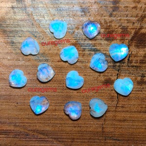 May include: A collection of heart-shaped moonstone gemstones. The stones display a range of blue and white hues, with a shimmering, iridescent quality. The gemstones are arranged on a wooden surface. The text "ouregems" is visible on the image.
