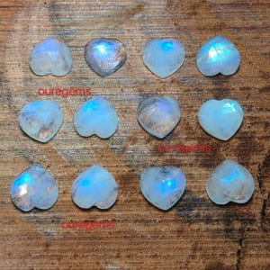 May include: A collection of heart-shaped moonstone gemstones. The stones are a translucent white with blue and iridescent flashes. The gemstones are arranged on a wooden surface. The text "puregems" is visible in the image.