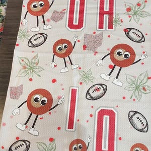 May include: A light gray towel featuring the letters "OH IO" in red, along with cartoon buckeye characters, footballs, and leaf designs. The towel has a repeating pattern of these elements.
