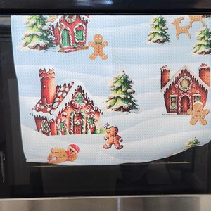 Op de afbeelding: Een witte keukendoek met een winterse scène. Het toont peperkoekhuizen, peperkoekmannetjes en kerstbomen. De huizen zijn rood met witte glazuur en snoepdetails. De doek hangt aan een ovendeur.
