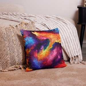 May include: A square throw pillow featuring a vibrant nebula print with swirling colors of deep blue, bright orange, magenta, and yellow. The design depicts a cosmic cloud of gas and dust with scattered white stars against a dark background.