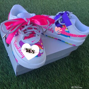 May include: White sneakers with pink and purple accents, rainbow designs, and the name "Enislaay" on the side. The shoes feature pink and purple ribbon laces and sit on a white box on a green surface.