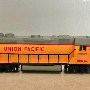Puede incluir: Una locomotora de tren a escala en amarillo y gris con barandillas negras. El lateral presenta el texto rojo UNION PACIFIC y el número 866. La máquina tiene un techo gris, ruedas negras y una cabina detallada con ventanas laterales.