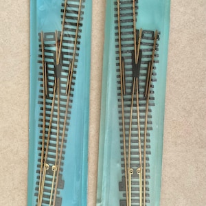 Peut inclure: Deux aiguillages de modélisme ferroviaire, étiquetés #6 Left et #6 Right, de la série Atlas Custom Line. Les rails sont noirs avec des rails couleur laiton, emballés dans du plastique transparent avec un support bleu. L'emballage contient du texte.