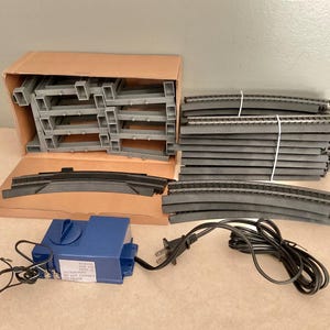 May include: A model train set including a blue power pack with a black power cord, gray plastic track supports, and curved black track sections. The power pack features a white label with black text that reads REMOVE FOR USE WITH AC ACCESSORIES DO NOT CONNECT TO TRACK.