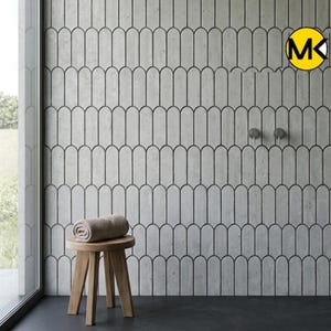 Op de afbeelding: Een badkamerinterieur met een lichtgrijze muur met een herhalend patroon van langwerpige, boogvormige vormen omlijnd in zwart. Een kleine houten kruk ondersteunt een opgerolde beige handdoek. Twee zilveren armaturen zijn aan de muur bevestigd. De vloer is donkergrijs.