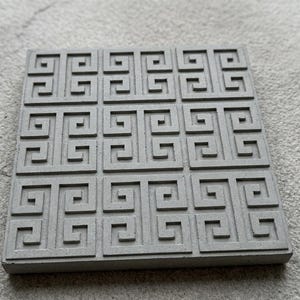 Op de afbeelding: Vierkante, grijze betonnen onderzetter met een herhalend Grieks sleutelpatroon. De onderzetter heeft een effen, neutrale kleur en een getextureerd oppervlak. Het ontwerp is een reeks in elkaar grijpende, geometrische vormen.