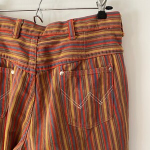K&ouml;nnte beinhalten: Eine gestreifte Hose mit hoher Taille und weitem Bein. Die Hose ist aus einem dicken, gewebten Stoff gefertigt und hat ein farbenfrohes Streifenmuster in Rot-, Orange-, Gelb-, Blau- und Braunt&ouml;nen.