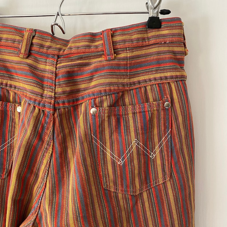 K&ouml;nnte beinhalten: Eine gestreifte Hose mit hoher Taille und weitem Bein. Die Hose ist aus einem dicken, gewebten Stoff gefertigt und hat ein farbenfrohes Streifenmuster in Rot-, Orange-, Gelb-, Blau- und Braunt&ouml;nen.