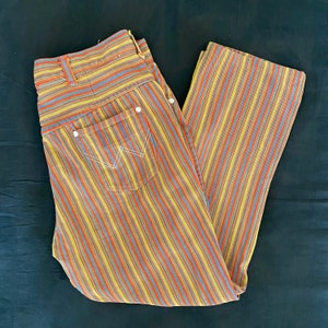 K&ouml;nnte beinhalten: Eine gestreifte Hose mit einem mehrfarbigen Muster aus Gelb, Orange, Rot, Blau und Grau. Die Hose hat eine vordere Tasche mit einem wei&szlig;en N&auml;hten-Detail.