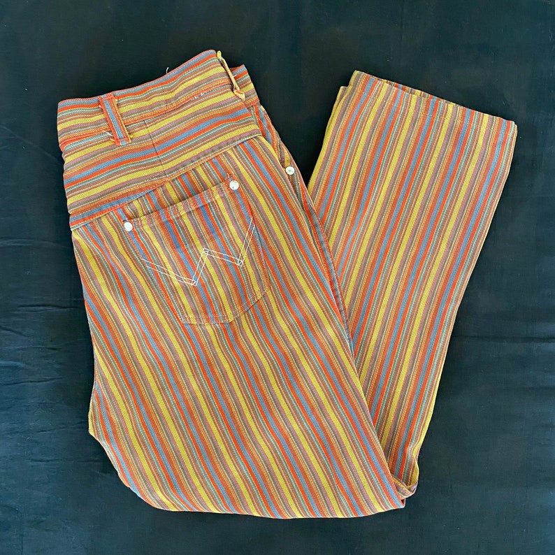 K&ouml;nnte beinhalten: Eine gestreifte Hose mit einem mehrfarbigen Muster aus Gelb, Orange, Rot, Blau und Grau. Die Hose hat eine vordere Tasche mit einem wei&szlig;en N&auml;hten-Detail.