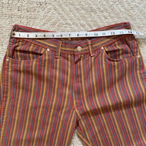 K&ouml;nnte beinhalten: Mehrfarbige gestreifte Hose mit hoher Taille und lockerer Passform. Die Hose hat ein rot-gelb-blau-braun gestreiftes Muster.