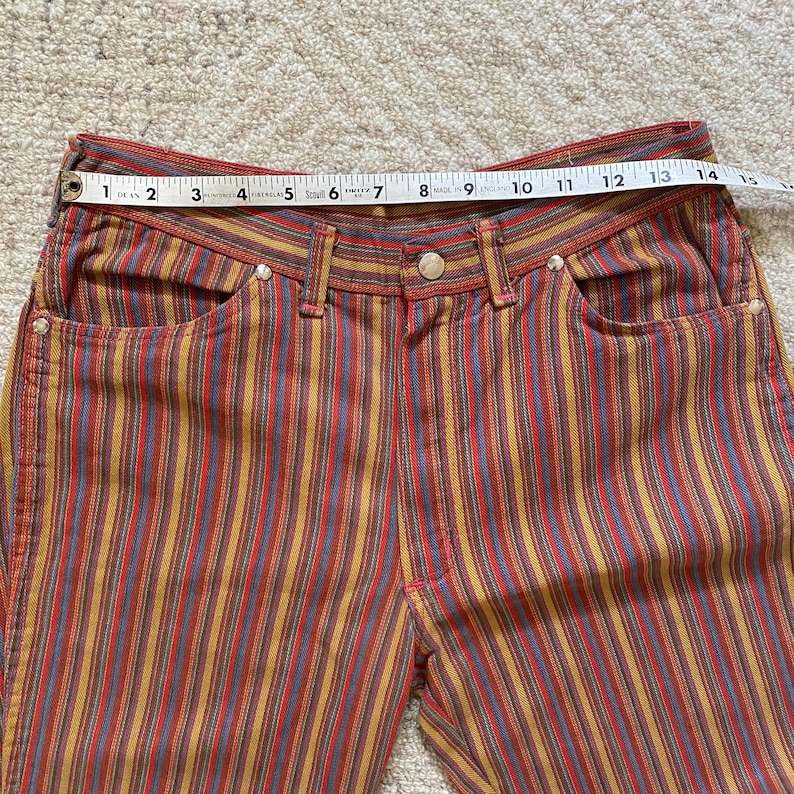 K&ouml;nnte beinhalten: Mehrfarbige gestreifte Hose mit hoher Taille und lockerer Passform. Die Hose hat ein rot-gelb-blau-braun gestreiftes Muster.