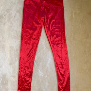 May include: A pair of vibrant red leggings featuring a smooth, shiny, and reflective finish. The garment has a high-waisted design with a simple elastic waistband and long, fitted legs that taper down to the ankles, showcasing a solid color.