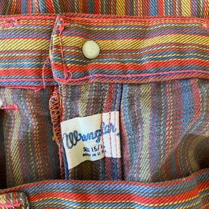 K&ouml;nnte beinhalten: Nahaufnahme eines Wrangler-Kleidungsetiketts, das an einer karierten Hose angen&auml;ht ist. Das Etikett ist wei&szlig; mit schwarzem Text und tr&auml;gt die Aufschrift "Wrangler" in einer stilisierten Schriftart. Das Etikett enth&auml;lt auch den Text "Gr&ouml;&szlig;e 15 1/2" und "Hergestellt in den USA". Die Hose ist kariert mit roten, blauen, braunen und gelben Streifen.