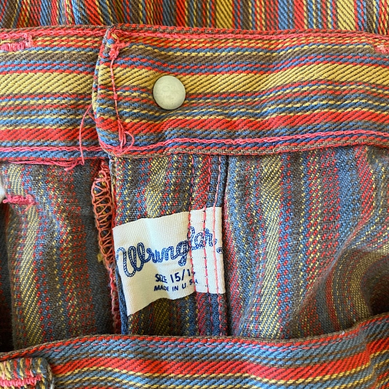 K&ouml;nnte beinhalten: Nahaufnahme eines Wrangler-Kleidungsetiketts, das an einer karierten Hose angen&auml;ht ist. Das Etikett ist wei&szlig; mit schwarzem Text und tr&auml;gt die Aufschrift "Wrangler" in einer stilisierten Schriftart. Das Etikett enth&auml;lt auch den Text "Gr&ouml;&szlig;e 15 1/2" und "Hergestellt in den USA". Die Hose ist kariert mit roten, blauen, braunen und gelben Streifen.