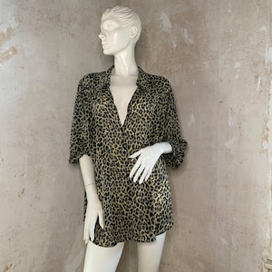 Puede incluir: Camisa transparente con estampado de leopardo, abotonada, expuesta en un maniquí. La camisa presenta un estampado negro y dorado, un cuello clásico y mangas tres cuartos. La prenda es ligera.