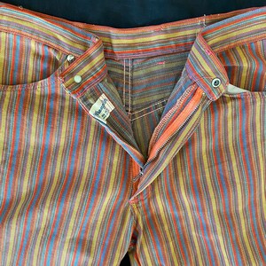K&ouml;nnte beinhalten: Eine gestreifte Wrangler-Jeans in einem rot, orange, gelb, blau und braunen Farbmuster. Die Jeans hat einen Rei&szlig;verschluss und ein Etikett mit der Aufschrift "Wrangler" und der Gr&ouml;&szlig;e "32 x 32".