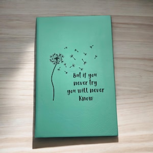 May include: A teal-colored journal with a dandelion illustration and the text "But if you never try you will never know." The dandelion is black and white, with seeds blowing away from the flower. The journal is rectangular and appears to be made of leather.