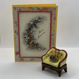 May include: A yellow and red porcelain trinket box shaped like a small armchair with a floral design. The box is closed and has a small gray cat figurine sitting on the seat. The box is sitting in front of a yellow card with a floral design and the text "Heureux Anniversaire".