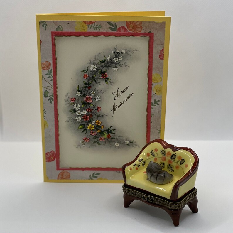 May include: A yellow and red porcelain trinket box shaped like a small armchair with a floral design. The box is closed and has a small gray cat figurine sitting on the seat. The box is sitting in front of a yellow card with a floral design and the text "Heureux Anniversaire".