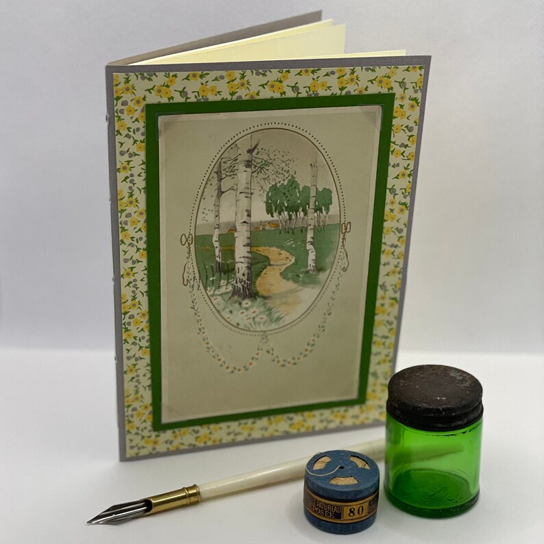 Puede incluir: Un diario con estampado verde y amarillo con una imagen de un camino a trav&eacute;s de un bosque de abedules. El diario est&aacute; cerrado y tiene un lomo gris. Un bol&iacute;grafo y una bobina de hilo est&aacute;n en primer plano.
