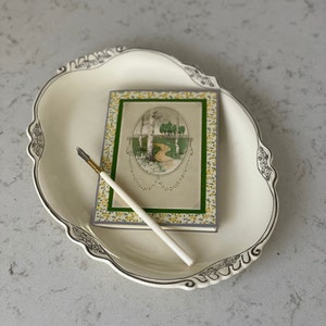 Puede incluir: Una fuente ovalada blanca con un borde decorativo y una postal vintage con un borde floral y una imagen de un camino a trav&eacute;s de un bosque. Un bol&iacute;grafo blanco y dorado est&aacute; sobre la fuente.