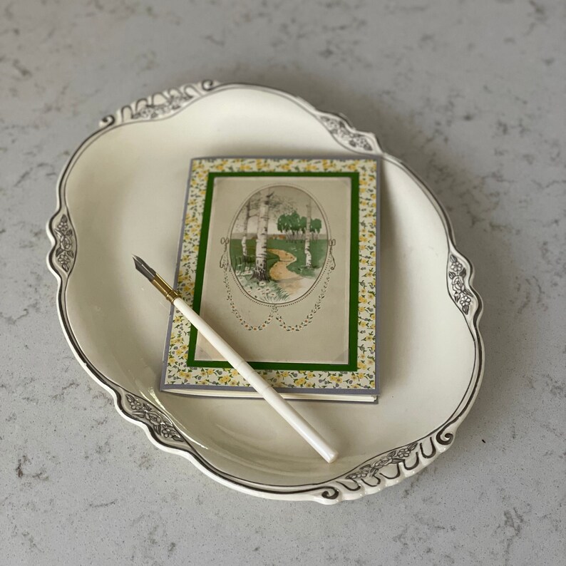 Puede incluir: Una fuente ovalada blanca con un borde decorativo y una postal vintage con un borde floral y una imagen de un camino a trav&eacute;s de un bosque. Un bol&iacute;grafo blanco y dorado est&aacute; sobre la fuente.