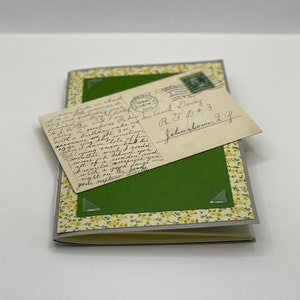 Puede incluir: Una postal vintage con texto escrito a mano y un sello verde. La postal est&aacute; pegada a un diario verde con un borde floral amarillo.