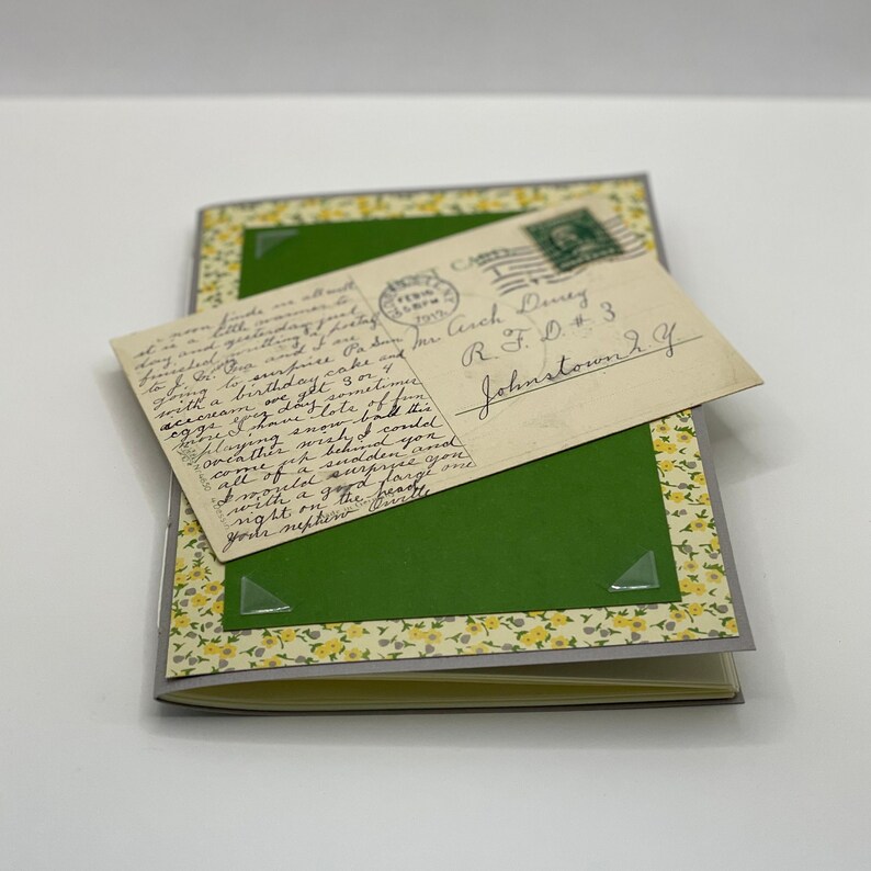 Puede incluir: Una postal vintage con texto escrito a mano y un sello verde. La postal est&aacute; pegada a un diario verde con un borde floral amarillo.
