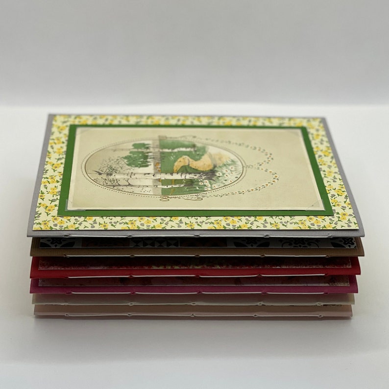 Puede incluir: Una pila de tarjetas hechas a mano con un dise&ntilde;o floral y una ilustraci&oacute;n vintage de un p&aacute;jaro en un jard&iacute;n. Las tarjetas est&aacute;n superpuestas en tonos de gris, marr&oacute;n, rojo, rosa y beige.