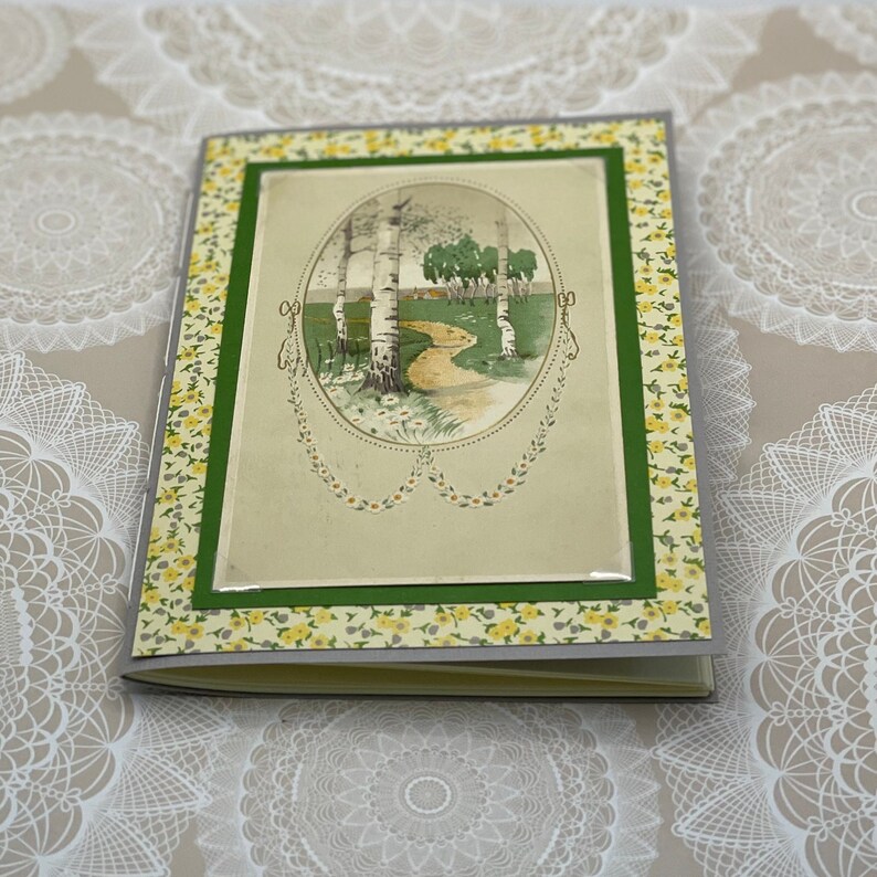 Puede incluir: Una tarjeta de estilo vintage con un borde floral verde y amarillo. La tarjeta presenta una imagen de un camino a trav&eacute;s de un bosque con abedules y una peque&ntilde;a casa en la distancia.
