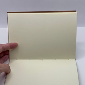 Puede incluir: Un cuaderno en blanco de color crema con una cubierta marr&oacute;n. El cuaderno est&aacute; abierto y las p&aacute;ginas est&aacute;n cosidas juntas con hilo blanco.