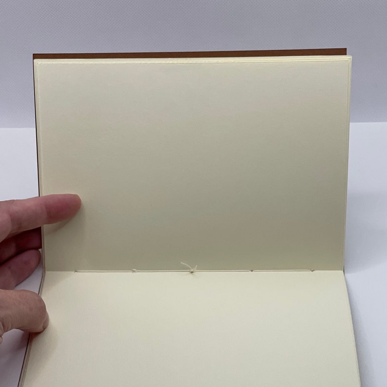 Puede incluir: Un cuaderno en blanco de color crema con una cubierta marr&oacute;n. El cuaderno est&aacute; abierto y las p&aacute;ginas est&aacute;n cosidas juntas con hilo blanco.