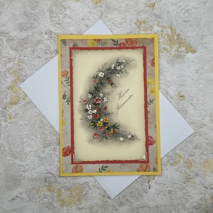 May include: A greeting card with a yellow border and a red inner border. The card features a floral design with a crescent moon shape. The text on the card reads "heureux anniversaire".