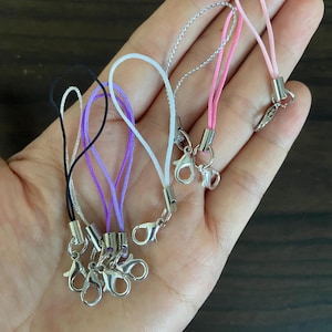 May include: A collection of colorful phone charms with silver-tone clasps. The charms feature cords in black, white, purple, pink, and silver. Each charm has a small metal clasp for easy attachment to a phone or other device.